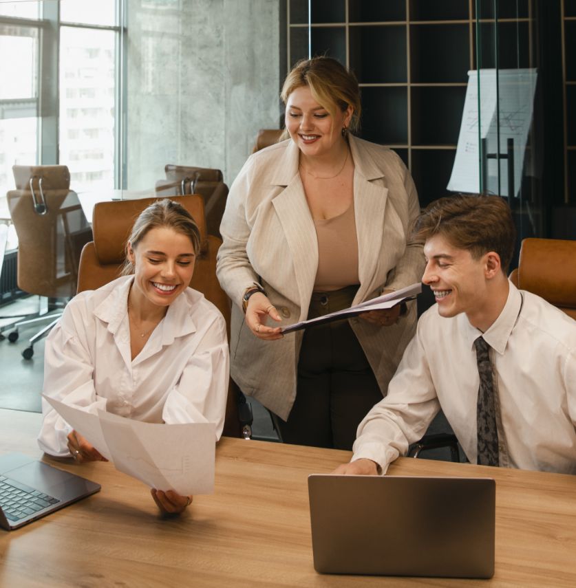 Three young professionals sit and stand around a wooden table in a modern office, smiling and reviewing documents together. Laptops and papers are spread out as they collaborate.