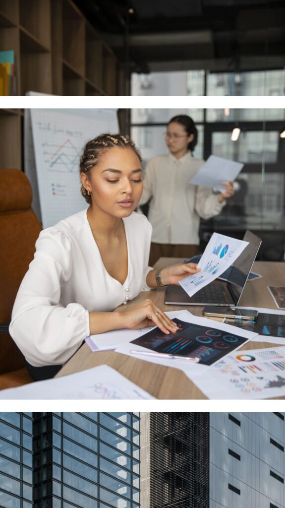 A woman sits at a desk reviewing charts and graphs in a modern office, while another stands in the background holding papers; a large window shows a city building outside.