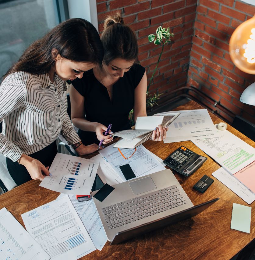 Two women work together at a wooden desk covered with papers, charts, a laptop, a calculator, and stationery in a modern office with a brick wall. They are focused on reviewing documents and financial data.