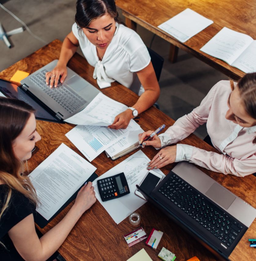 Three women sit at a wooden table working together on documents and laptops, with papers, a calculator, pens, and office supplies spread out in a collaborative workspace.