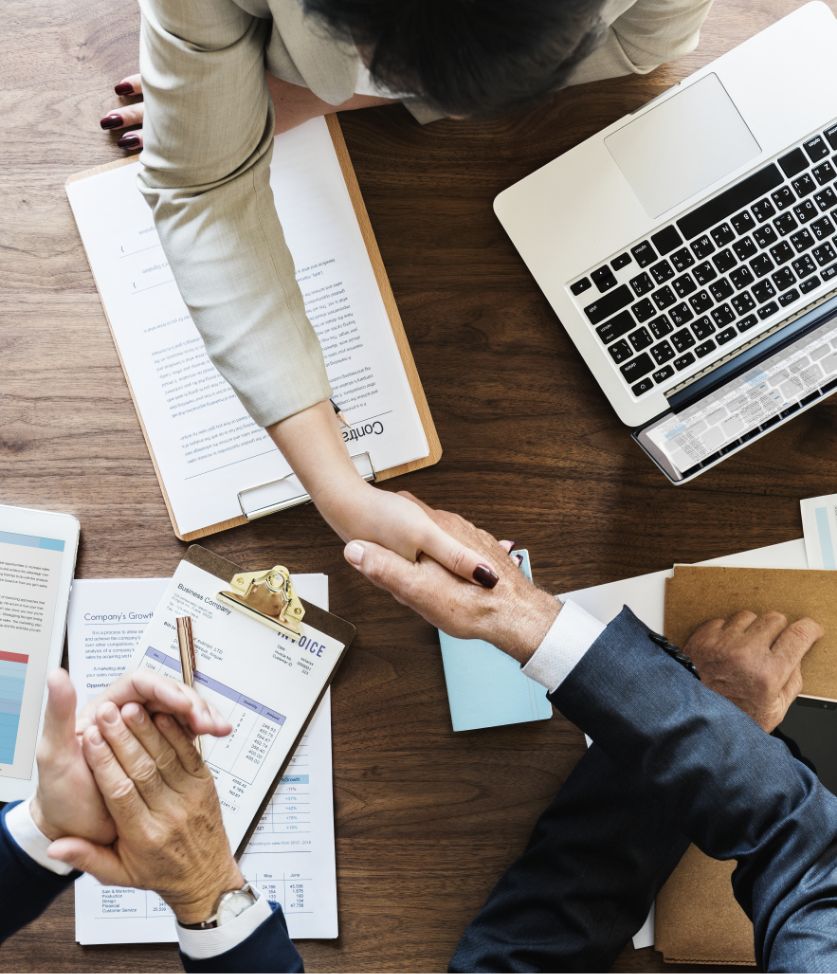 Two people in business attire shaking hands across a wooden desk with documents, charts, folders, and a laptop, symbolizing agreement or partnership in a professional setting.