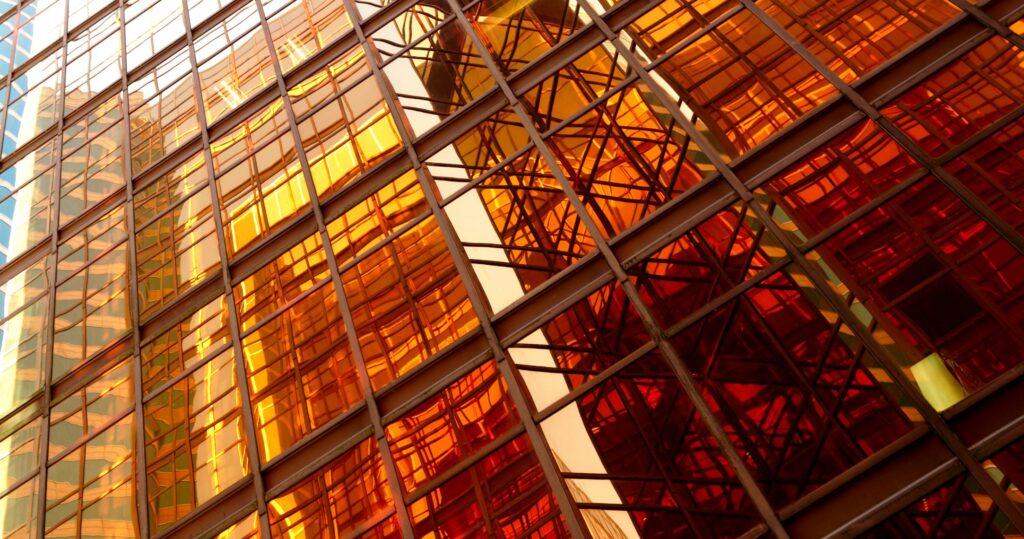 Close-up of a modern building facade with reflective glass panels in warm shades of orange, yellow, and red, showing abstract reflections of structural beams and the surrounding architecture.